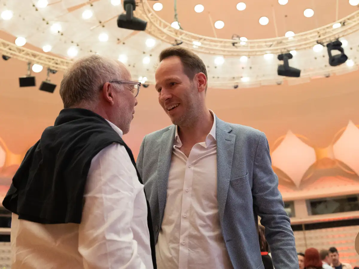 Zwei Männer unterhalten sich lächelnd in einem hell erleuchteten Raum mit runder Decke und vielen Lichtern.