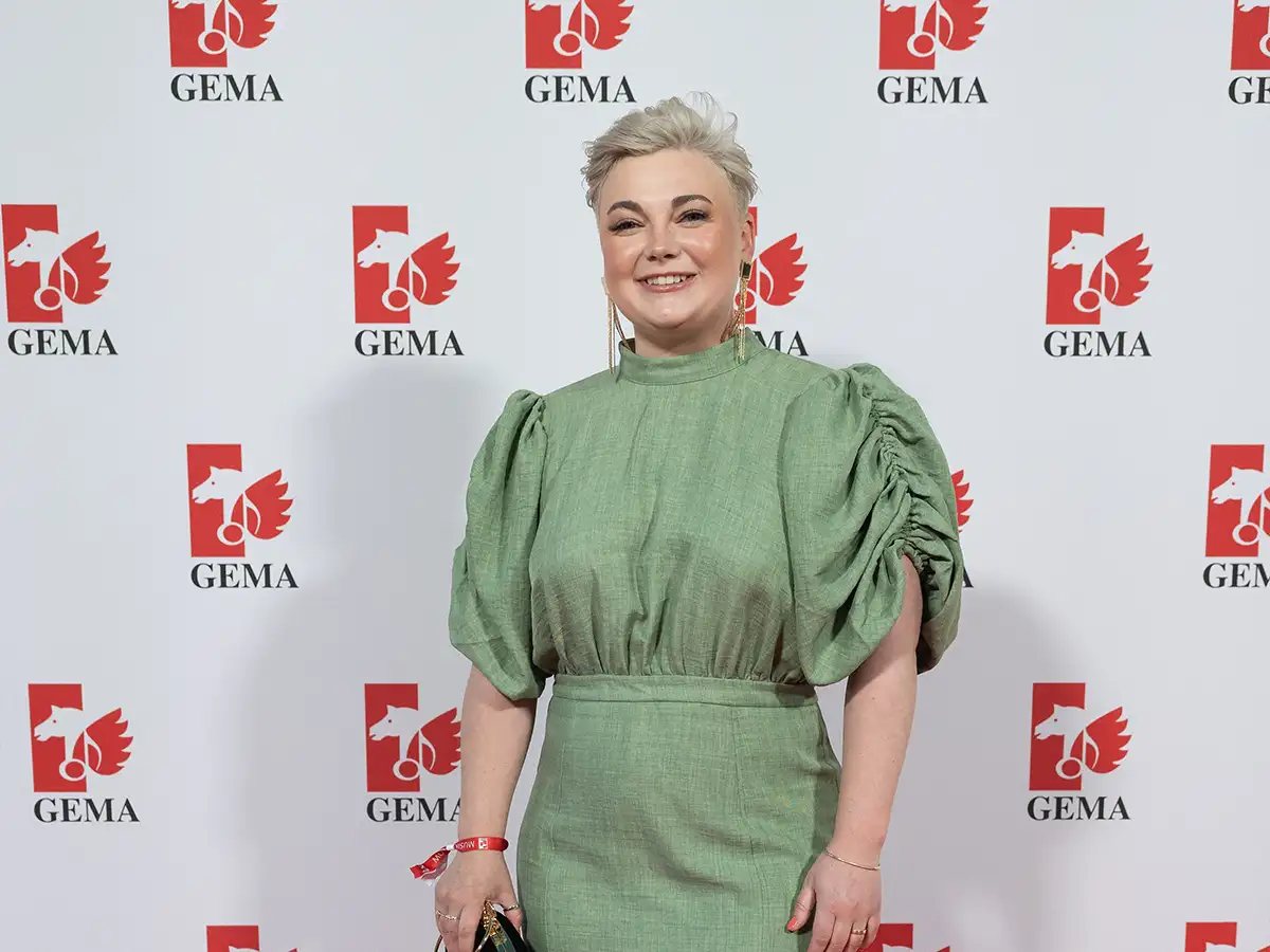 Eine Person in grünem Kleid steht vor einer GEMA-Wand und lächelt in die Kamera.