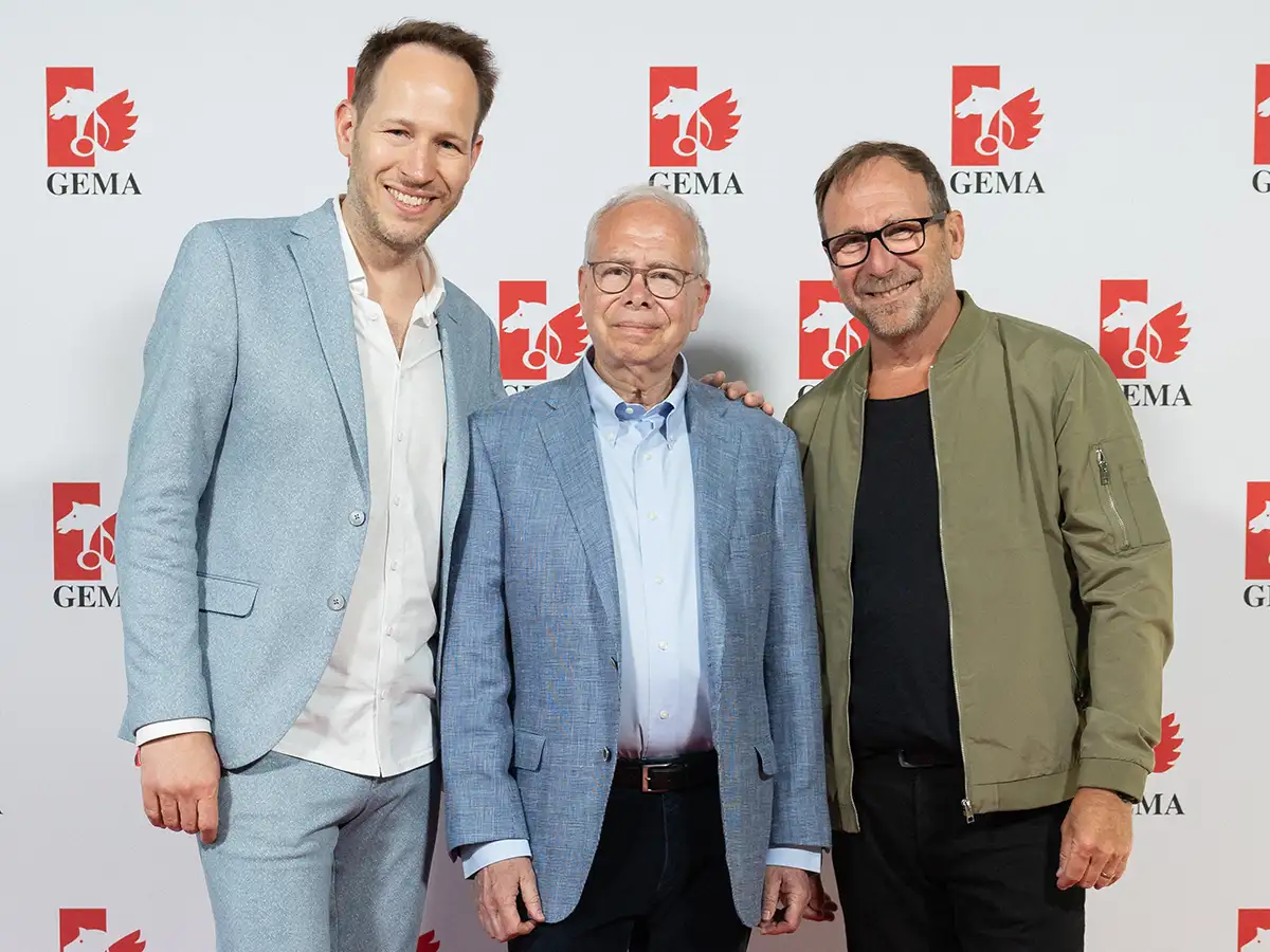 Drei Männer in Anzügen und Jacke stehen vor einer GEMA-Wand und lächeln in die Kamera.