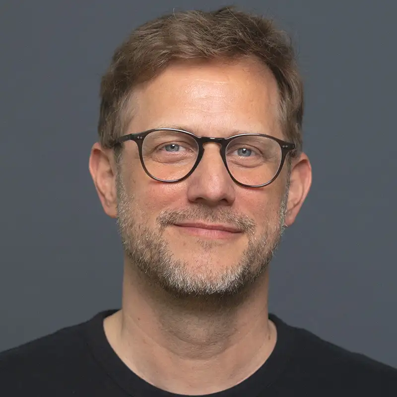 Ein lächelnder Mann mit Brille vor grauem Hintergrund.