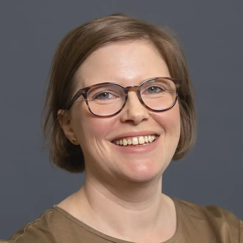 Eine lächelnde Person mit Brille und braunem Haar vor grauem Hintergrund.
