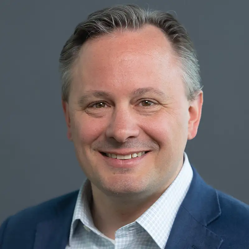 Ein lächelnder Mann in einem blauen Anzug vor grauem Hintergrund.