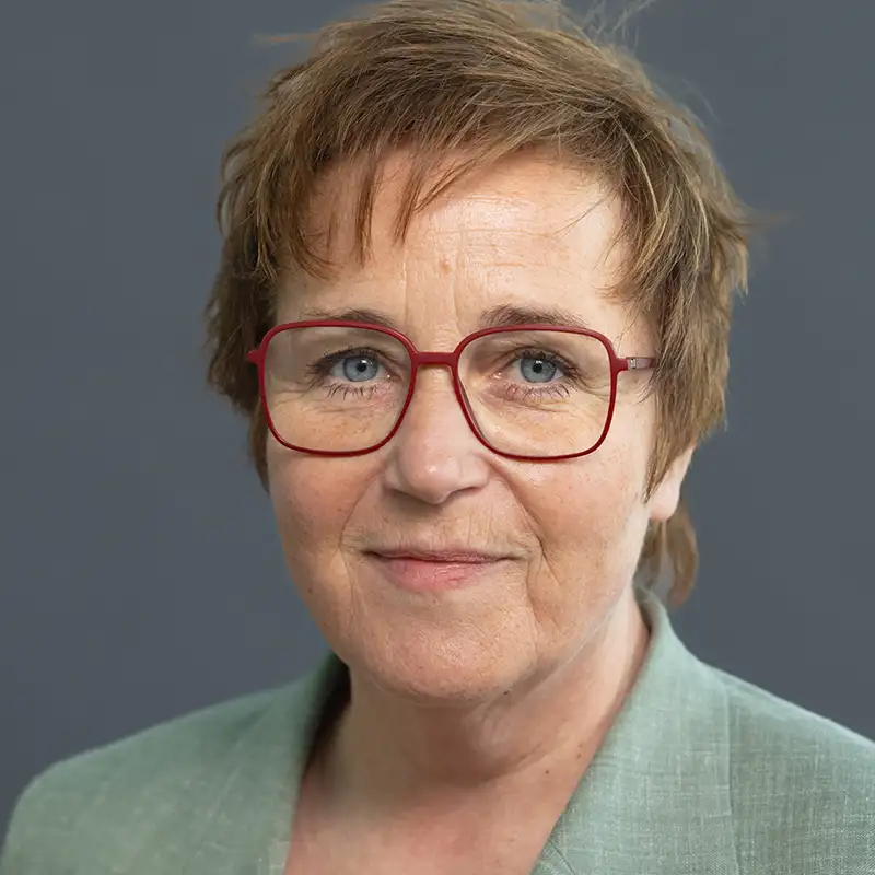 Eine Person mit kurzen Haaren und roter Brille vor grauem Hintergrund. Sie trägt ein grünes Jackett.