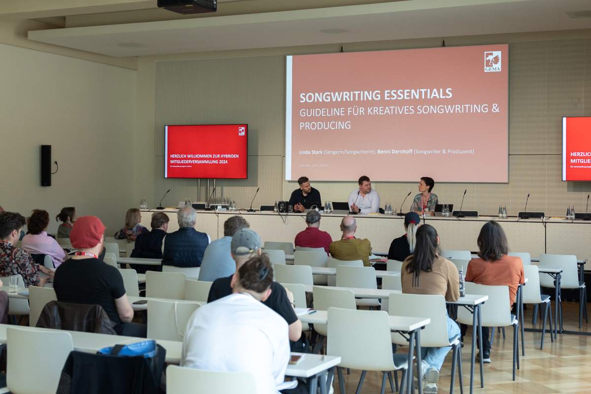 Ein Konferenzraum mit Publikum, drei Personen auf einem Podium, und einer Präsentation zu Songwriting an der Wand.