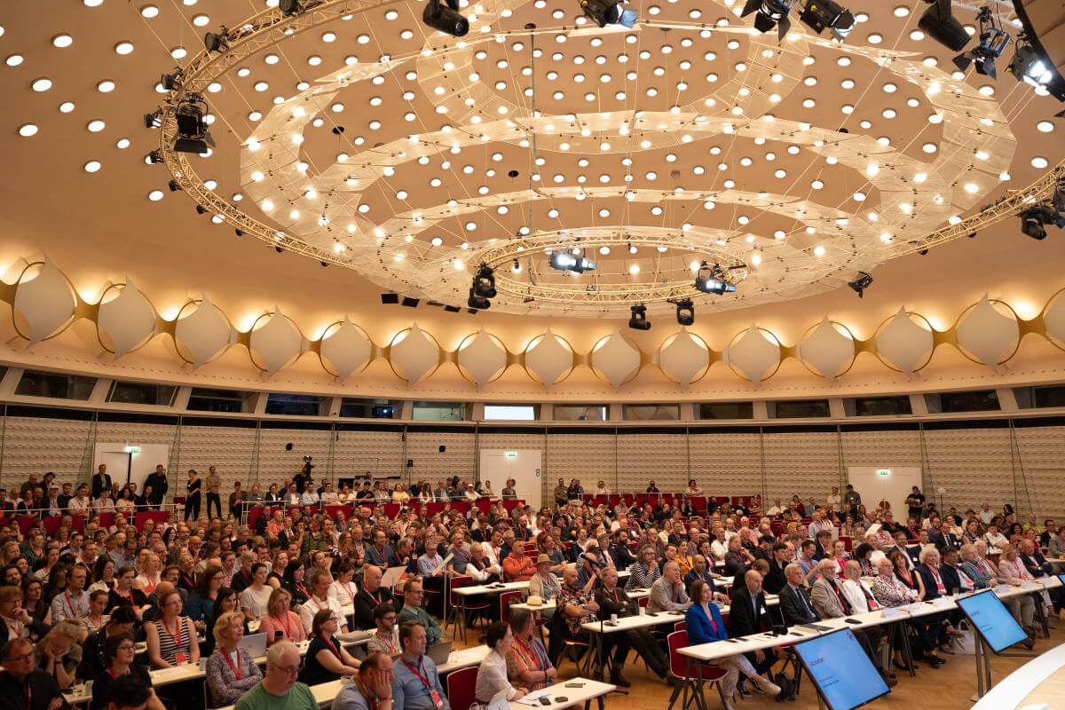 Großer Konferenzsaal mit vielen Menschen, runden Deckenleuchten und mehreren Bildschirmen.