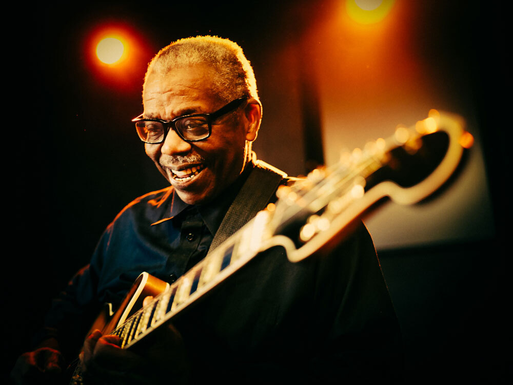 Ein lächelnder Mann spielt Gitarre unter warmem Licht.