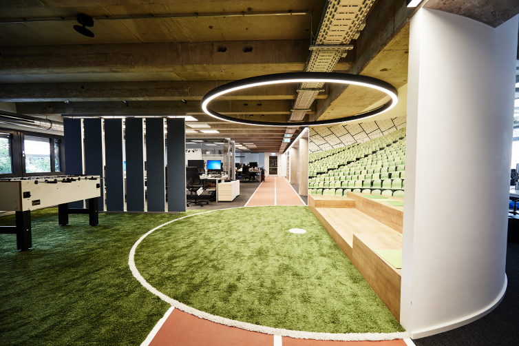 Ein modernes Büro mit Kunstrasen, Tribünen, Tischkicker und offenen Arbeitsplätzen unter einem großen, runden Deckenlicht.