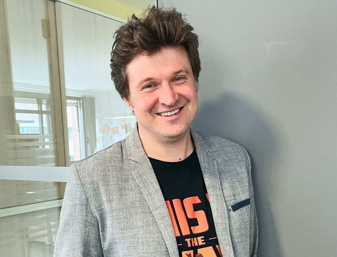 Ein Mann in grauem Sakko lächelt vor einer grauen Wand, im Hintergrund sind Glasfenster.