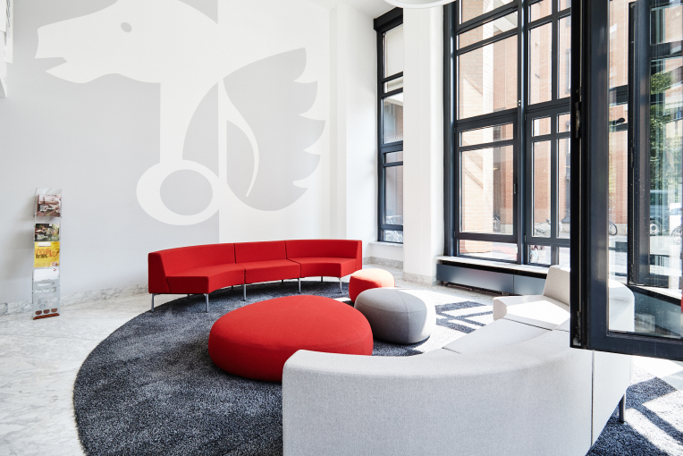 Ein moderner Wartebereich mit roten und grauen Sofas, runden Hockern und großen Fenstern, auf einem grauen Teppich.