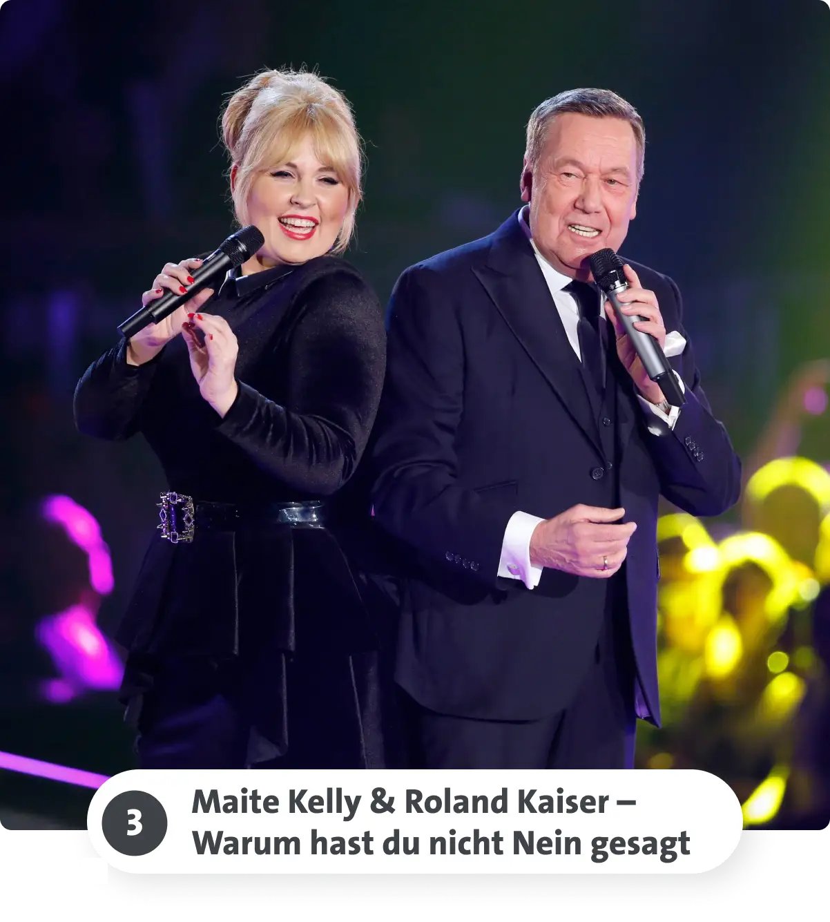 Zwei Personen singen auf einer Bühne, lächelnd und mit Mikrofonen in der Hand, vor einem bunten Hintergrund.