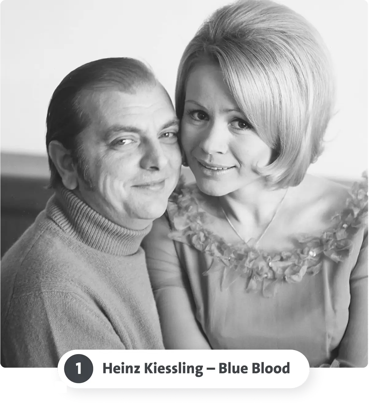 Ein lächelndes Paar in Schwarz-Weiß, sie trägt ein Kleid mit Blumenmuster, er einen Rollkragenpullover.