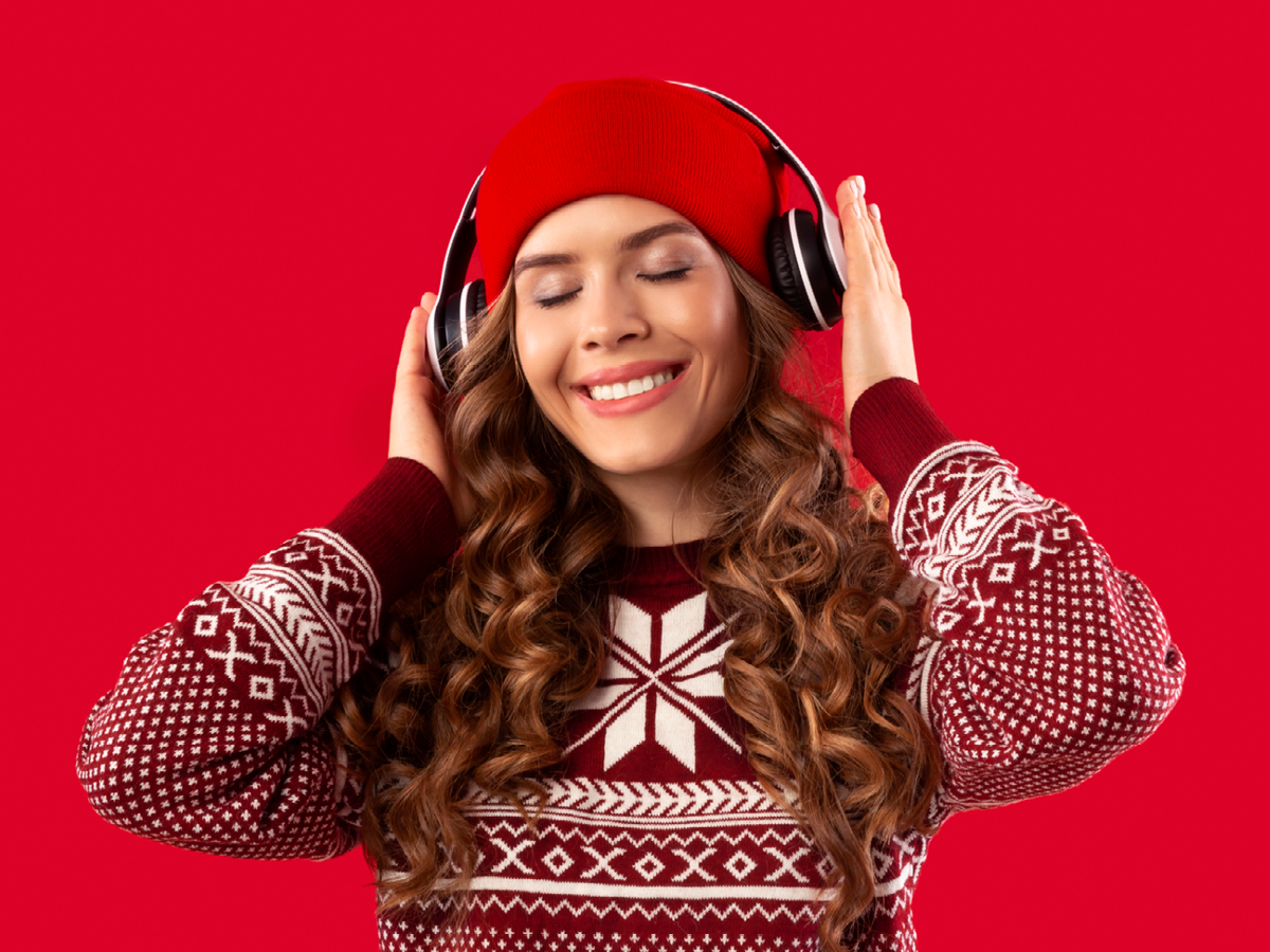 Frau mit roter Mütze und Kopfhörern, lächelnd, in einem rot-weißen Winterpullover vor rotem Hintergrund.