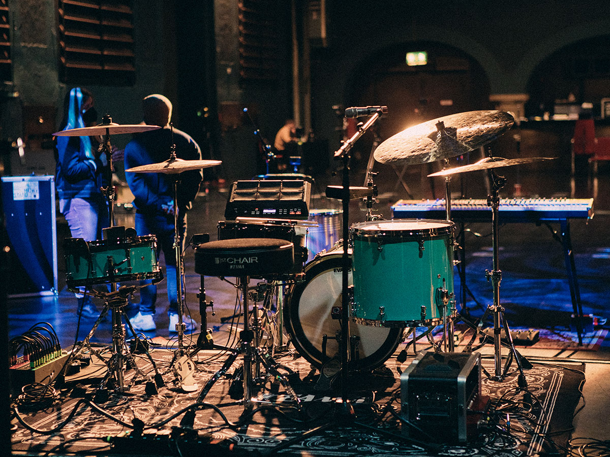 Ein Schlagzeug-Set auf einer Bühne, beleuchtet, mit zwei Personen im Hintergrund.