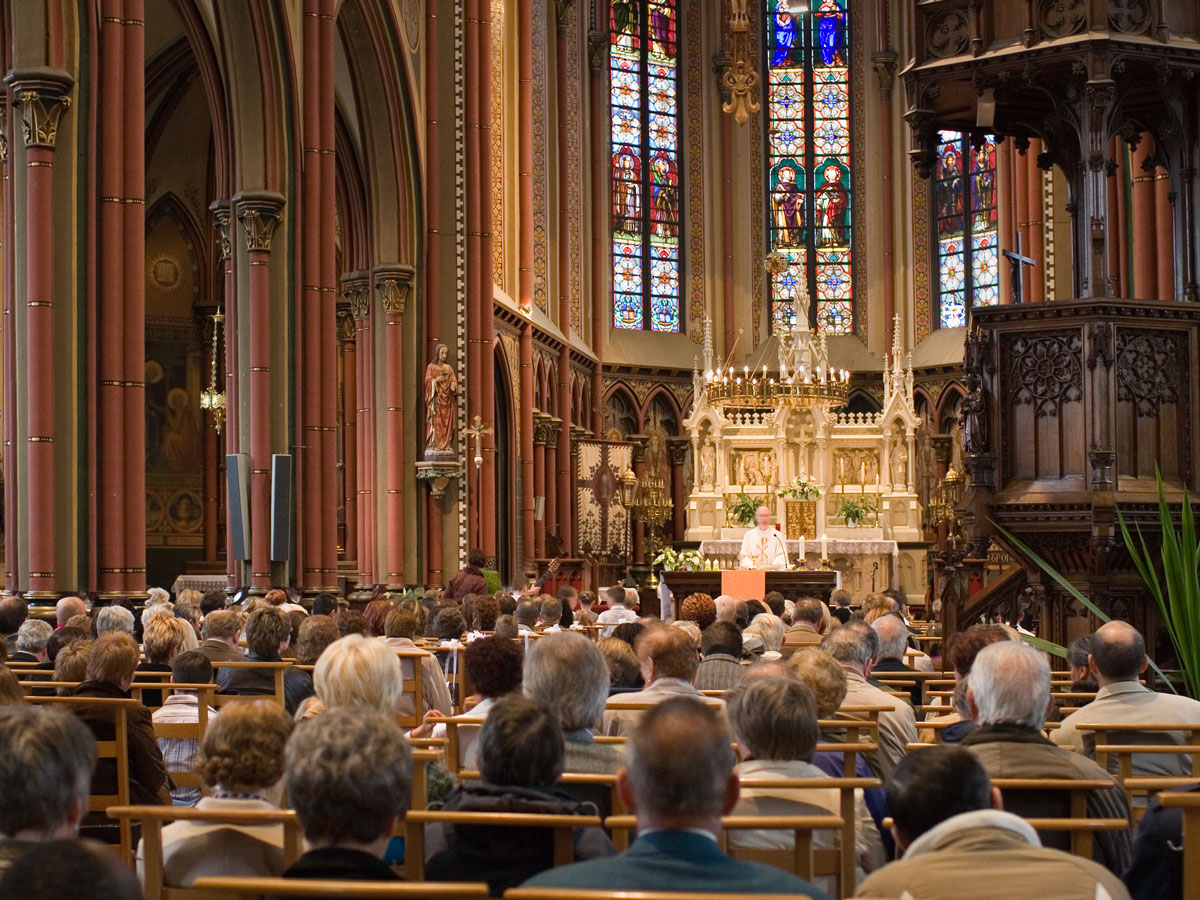 Eine volle Kirche mit Gläubigen, die einem Priester vor einem Altar mit bunten Glasfenstern zuhören.