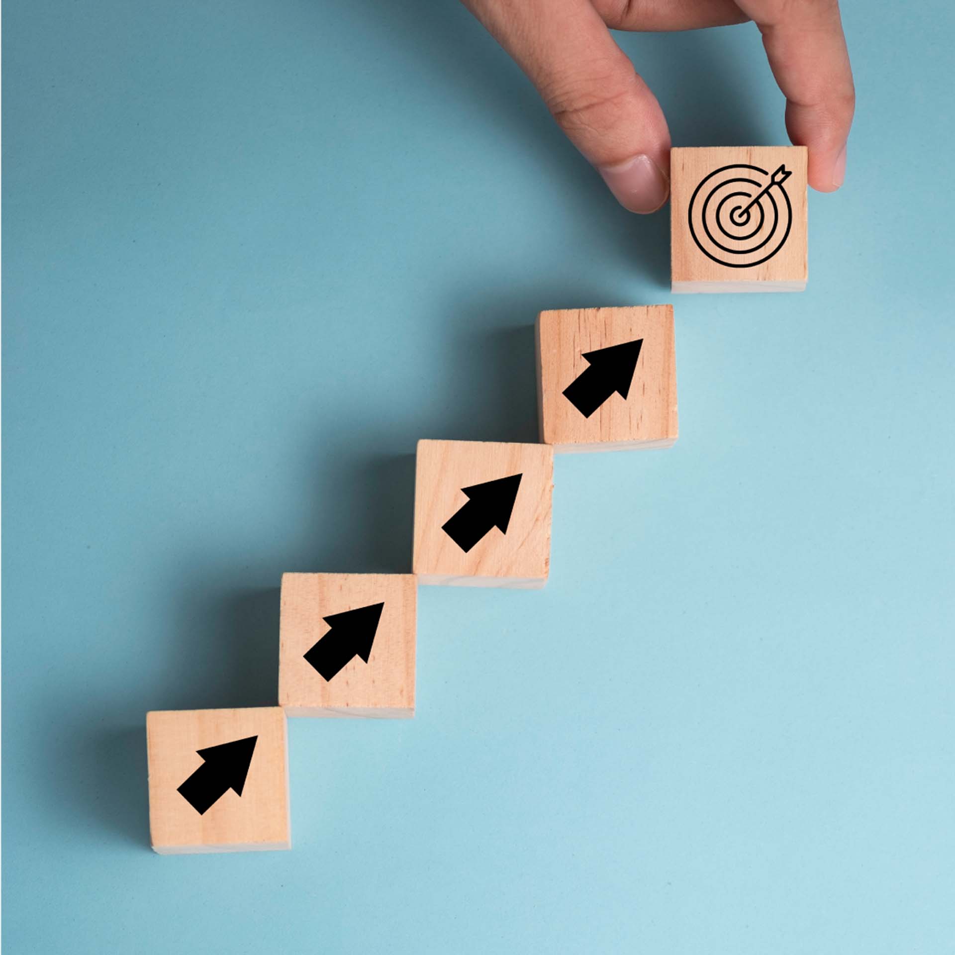 Holzwürfel mit Pfeilen bilden eine Treppe, oben ein Zielsymbol, auf hellblauem Hintergrund.