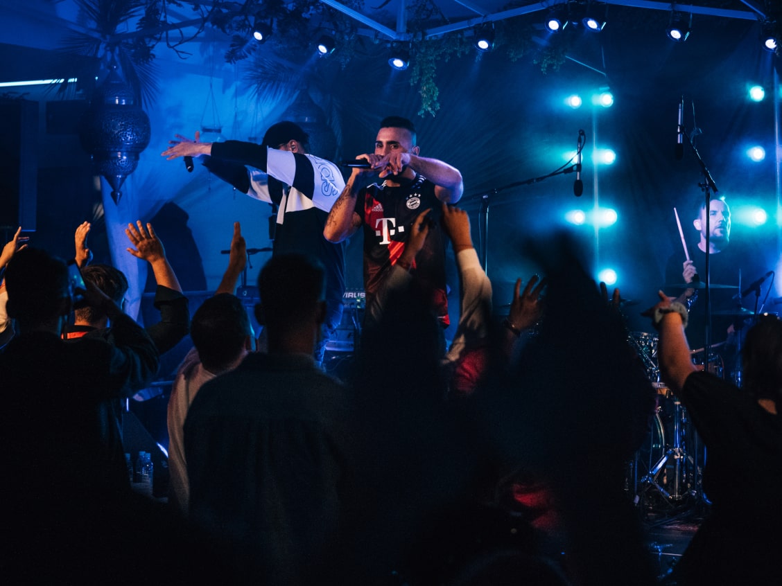 Ein Sänger und ein DJ performen auf einer Bühne mit blauer Beleuchtung, umgeben von einem begeisterten Publikum.