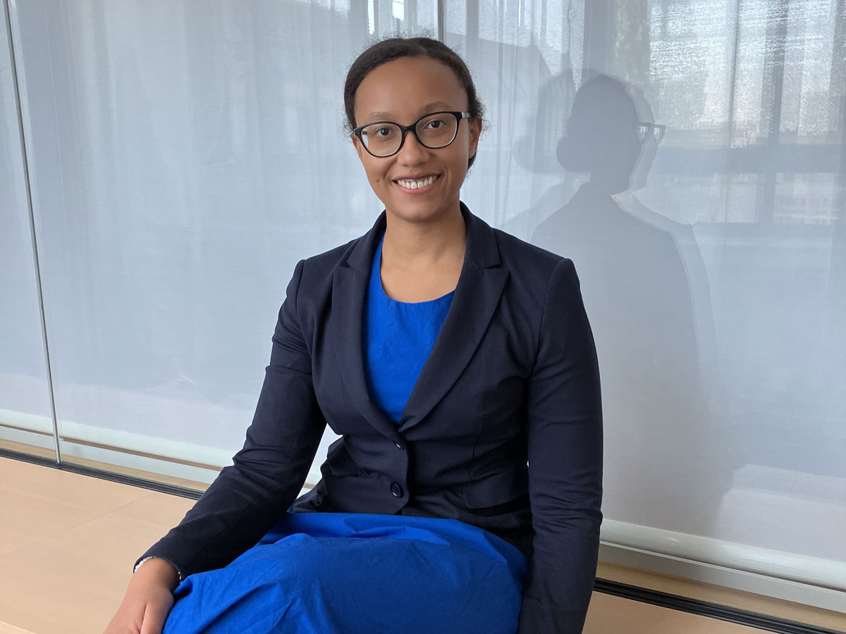 Eine Person in blauem Kleid und schwarzem Blazer sitzt lächelnd vor einem Fenster.