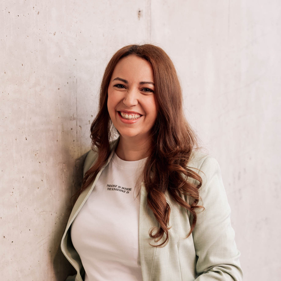 Eine lächelnde Frau mit braunen Haaren lehnt an einer Betonwand.