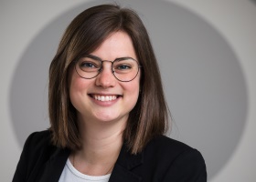 Eine lächelnde Person mit Brille und braunen Haaren vor grauem Hintergrund.