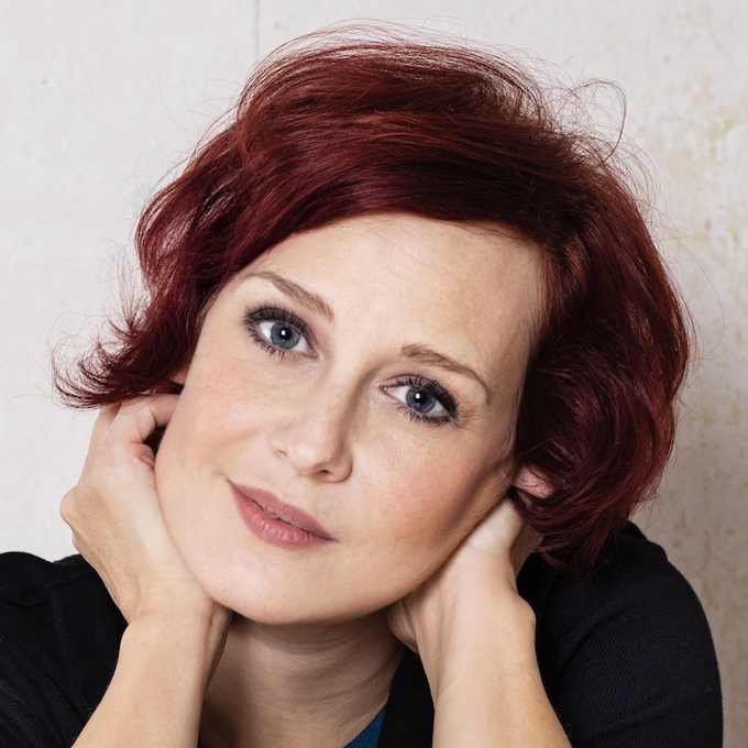 Eine Frau mit roten Haaren stützt ihr Gesicht in die Hände und blickt freundlich in die Kamera.