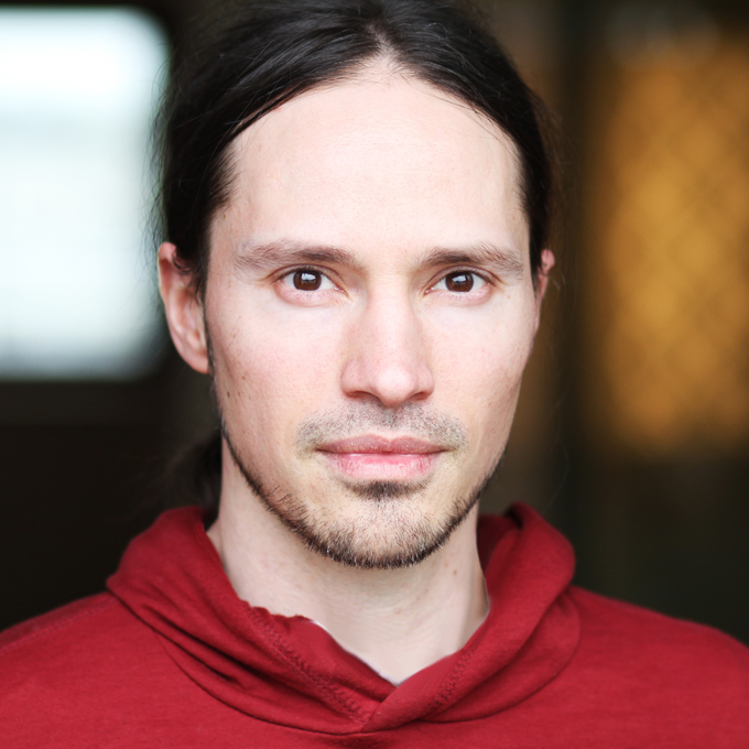 Eine Person mit langen Haaren trägt einen roten Pullover und blickt direkt in die Kamera.