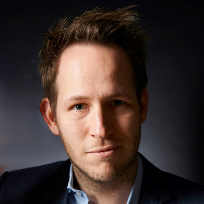 Ein Mann mit kurzen Haaren und einem leichten Lächeln, der eine dunkle Anzugsjacke und ein helles Hemd trägt, vor einem dunklen Hintergrund.