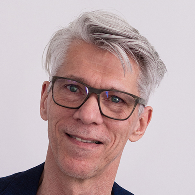 Ein Mann mit grauen Haaren und Brille lächelt vor neutralem Hintergrund.