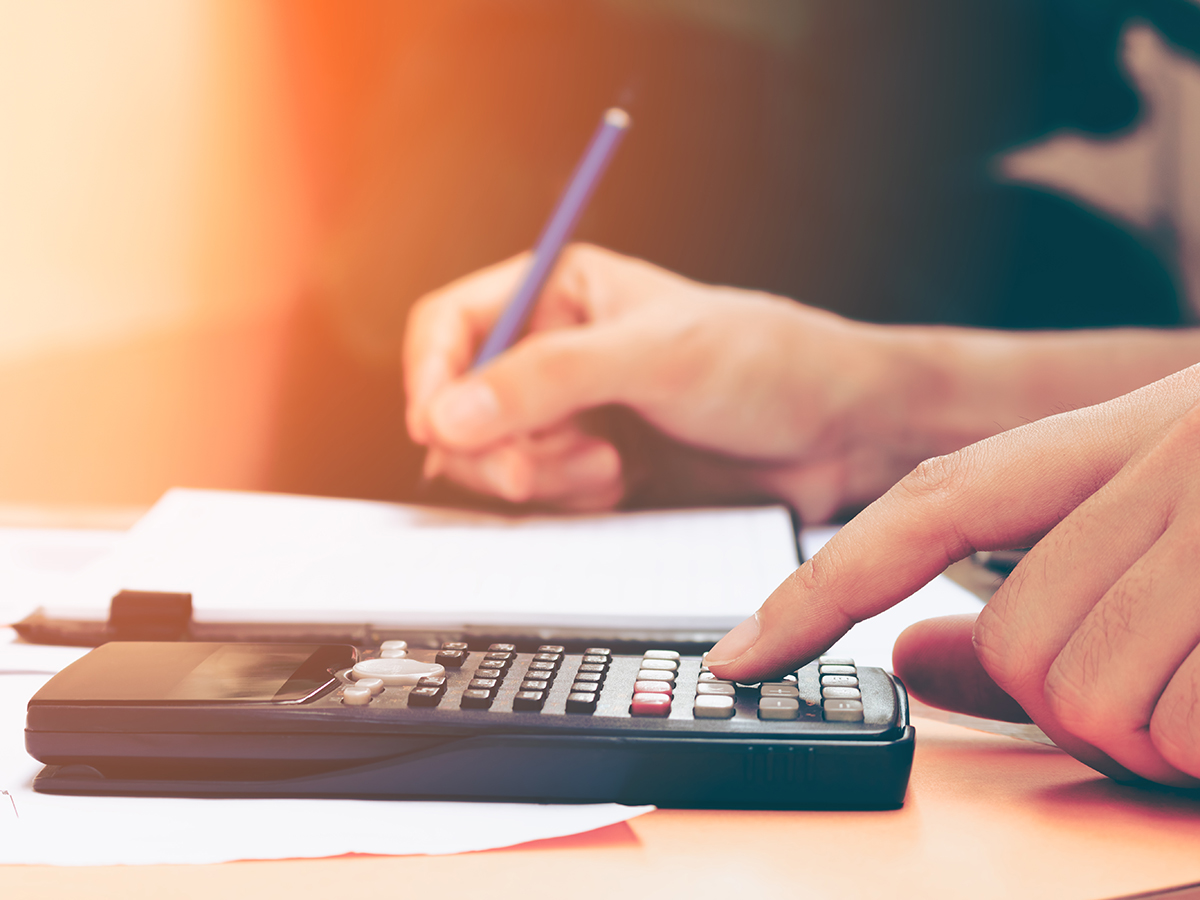 Hand tippt auf Taschenrechner, während eine andere Hand mit Stift auf Papier schreibt.