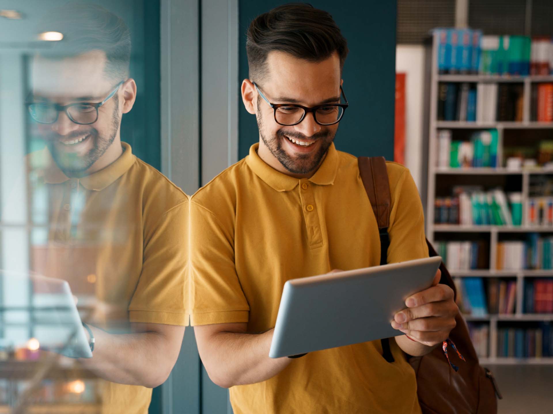 Ein Mann mit Brille und gelbem Hemd lächelt beim Tablet-Studium in einer Bibliothek.