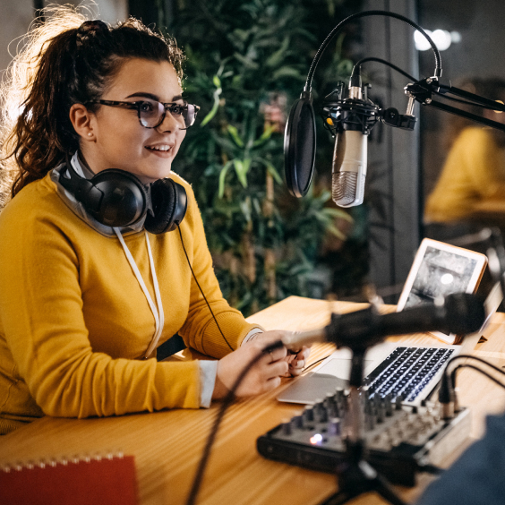 Eine Frau mit Brille und Kopfhörern spricht in ein Mikrofon vor einem Laptop.