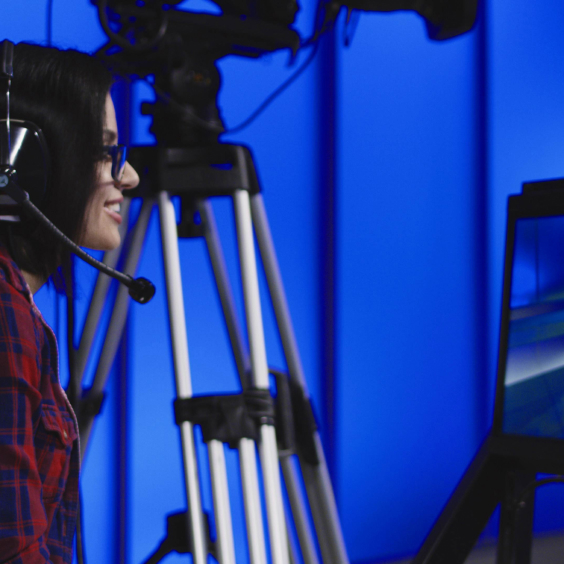 Frau mit Headset sitzt vor Monitoren, umgeben von Kamerastativen, vor blauem Hintergrund.