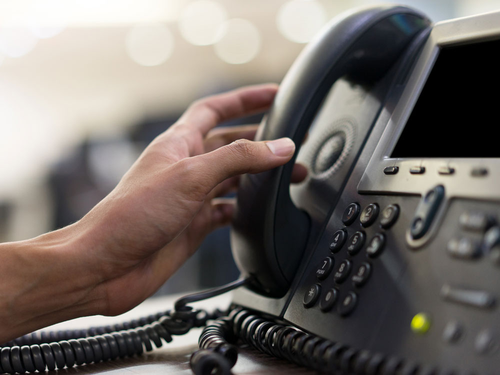 Eine Hand hebt den Hörer eines Bürotelefons ab.