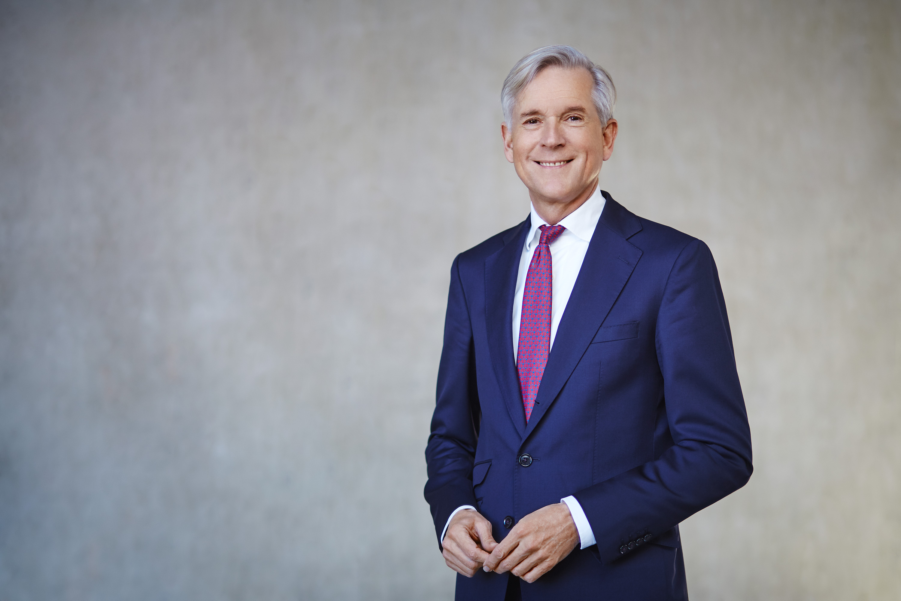 Ein lächelnder Mann in einem blauen Anzug mit roter Krawatte steht vor einer grauen Wand.