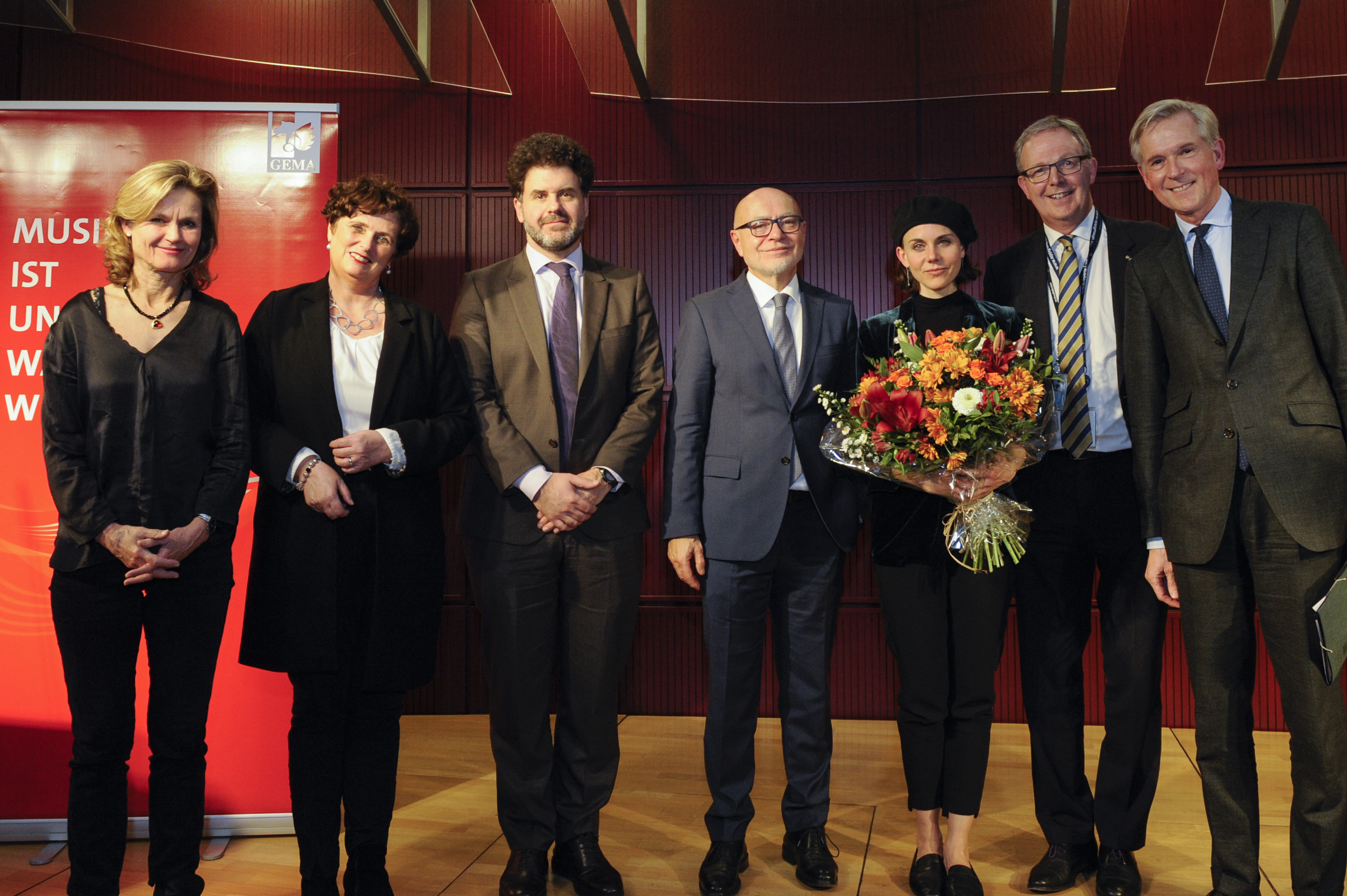 Sieben Personen stehen nebeneinander, eine Frau hält einen Blumenstrauß, rotes Banner im Hintergrund.