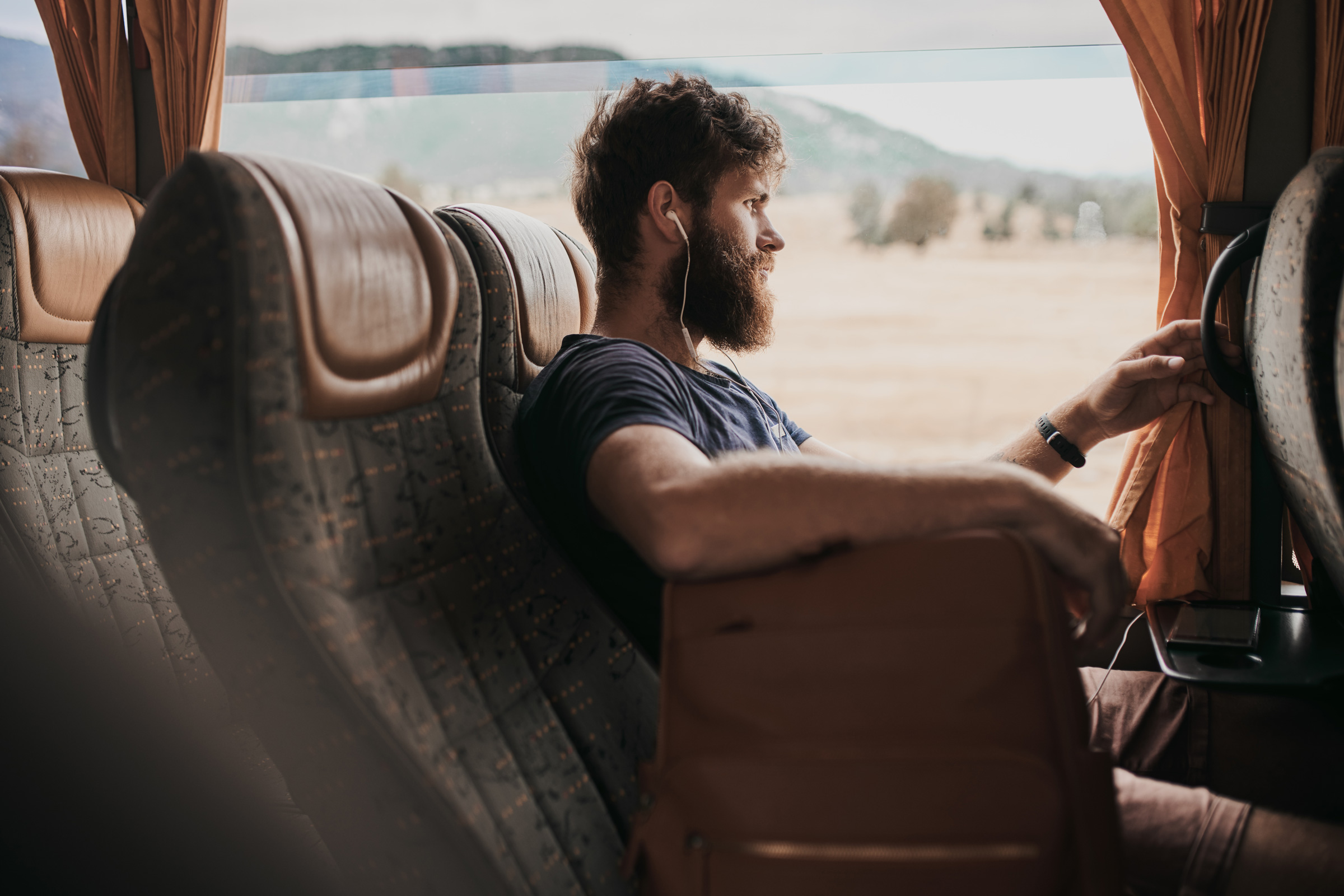 Ein Mann mit Bart sitzt im Bus, schaut aus dem Fenster, trägt Kopfhörer, umgeben von orangefarbenen Vorhängen.