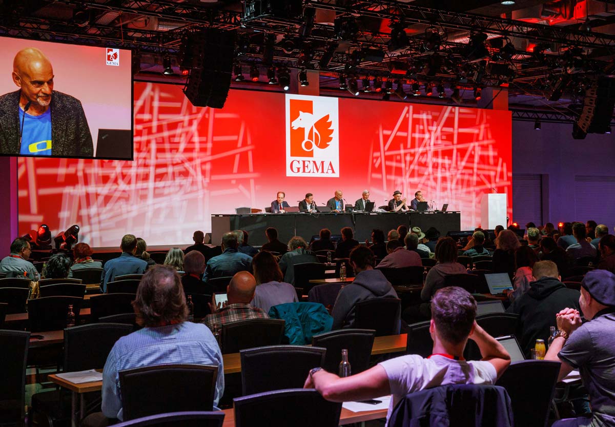 Menschen sitzen in einem Konferenzraum, vorne eine Bühne mit Rednern, großer Bildschirm zeigt eine Präsentation.