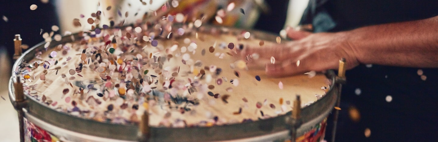 Eine Hand trommelt auf einer Trommel, während bunte Konfetti in die Luft fliegen.