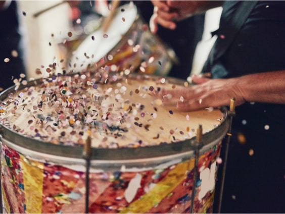 Bunte Konfetti fliegen von einer Trommel, die von einer Hand gespielt wird.