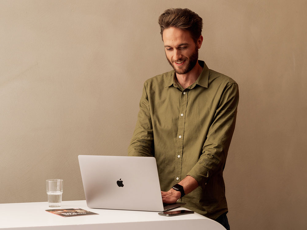 Ein Mann in grünem Hemd arbeitet lächelnd an einem Laptop auf einem Tisch mit Glas Wasser und Smartphone.