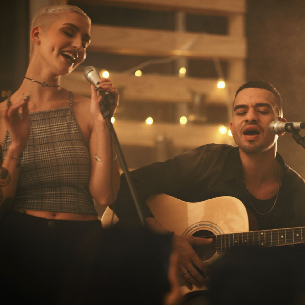 Zwei Personen singen und spielen Gitarre in gemütlicher, mit Lichtern dekorierter Umgebung.