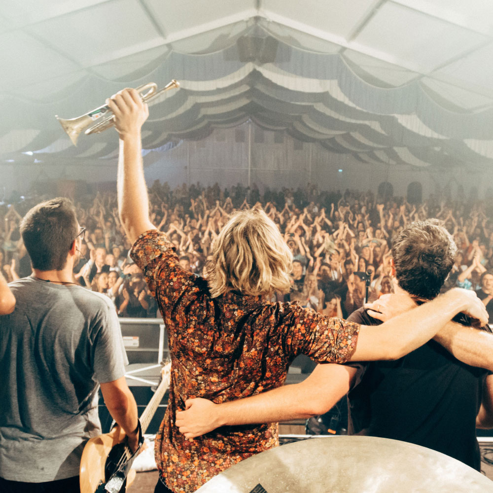 Drei Musiker stehen auf der Bühne, umarmen sich und blicken auf jubelndes Publikum.