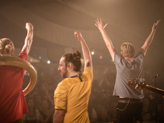 Drei Musiker auf der Bühne, Arme erhoben, Publikum im Hintergrund, verschwommene Lichter.