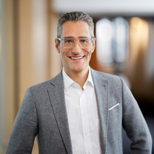 Ein Mann in grauem Anzug und weißem Hemd lächelt in die Kamera, unscharfer Hintergrund.