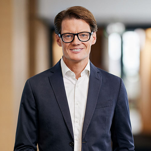 Ein Mann in Anzug und Brille lächelt in einem Büro mit verschwommenem Hintergrund.