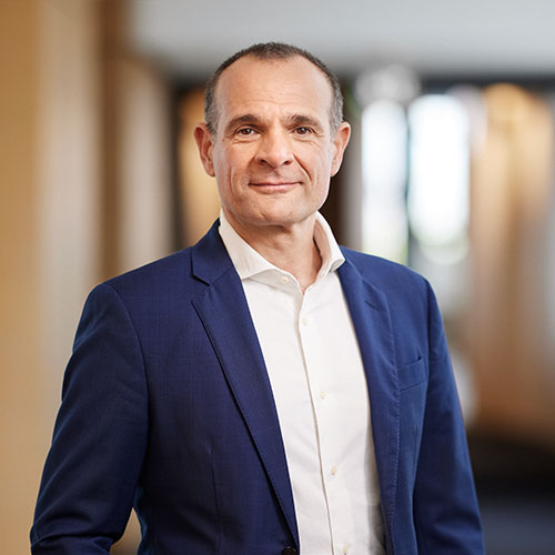 Ein Mann im blauen Anzug steht in einem hellen Raum mit verschwommenem Hintergrund.