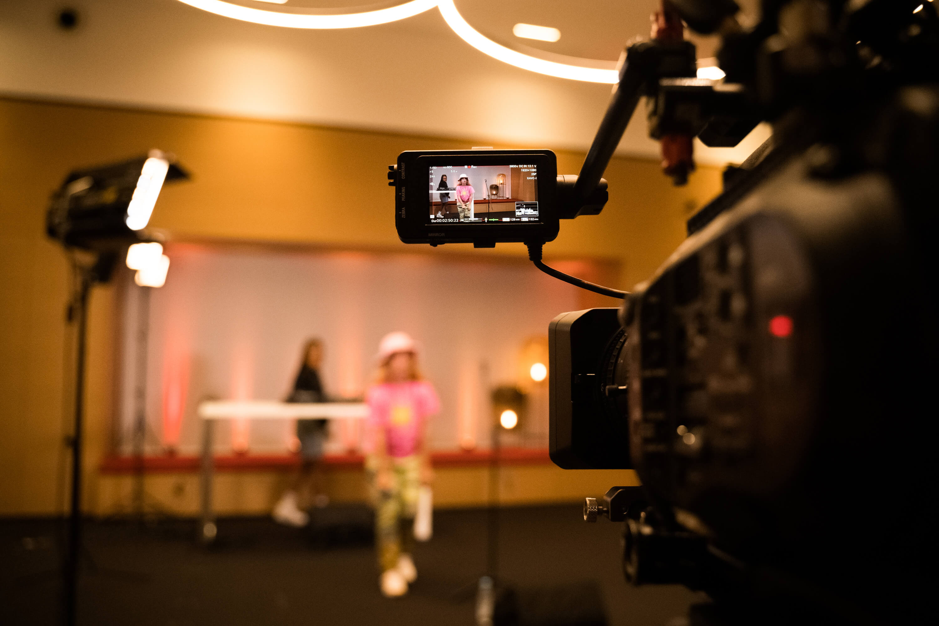 Kamera filmt Person in pinkem Outfit auf Bühne, unscharfer Hintergrund mit Lichtern.