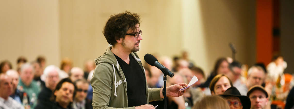 Ein Mann spricht in ein Mikrofon vor einem Publikum, hält dabei Notizen in der Hand.