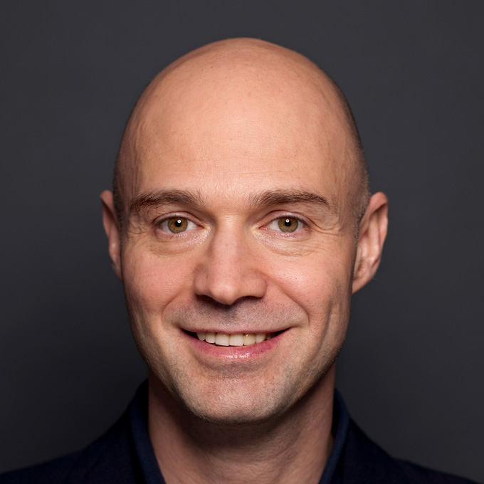 Ein lächelnder, glatzköpfiger Mann vor grauem Hintergrund.