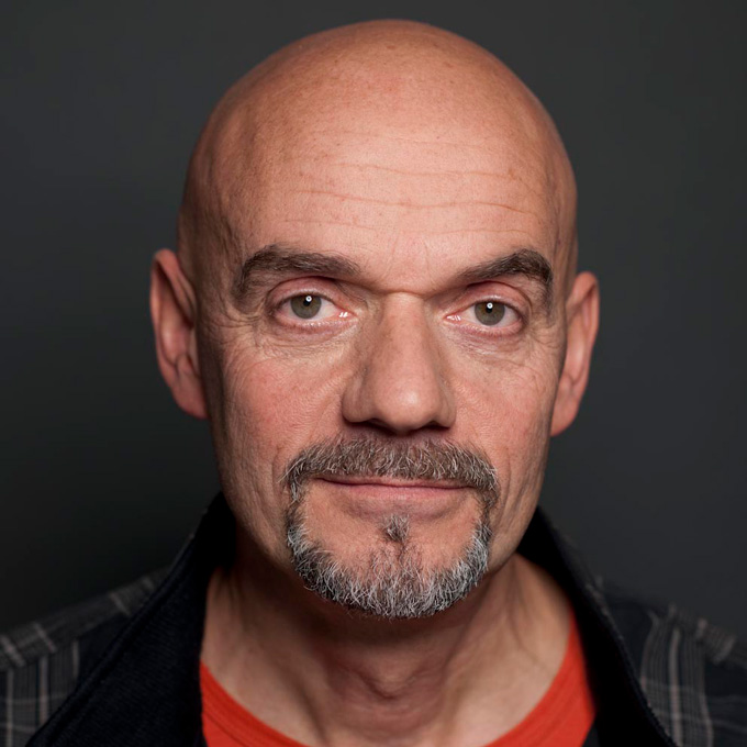 Ein Mann mit Glatze und Bart schaut direkt in die Kamera vor einem dunklen Hintergrund.