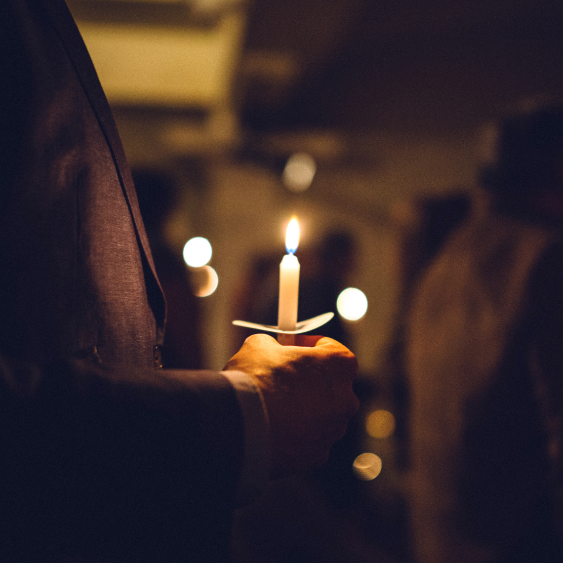 Eine Hand hält eine brennende Kerze in einem dunklen Raum, umgeben von unscharfen Lichtern.